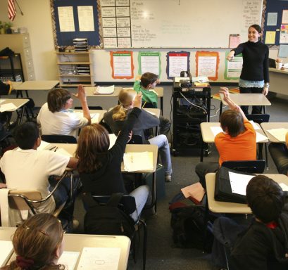 Estudiantes levantando la mano en un aula mientras un maestro expone un tema.
