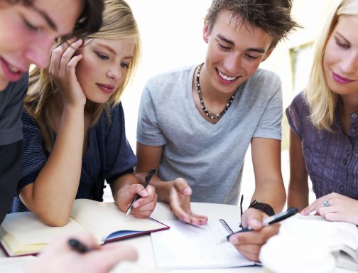 Grupo de cuatro jóvenes colaborando en un proyecto, escribiendo y tomando notas.