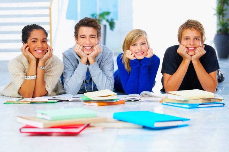 Cuatro jóvenes sonrientes sentados en el suelo con libros abiertos a su alrededor.