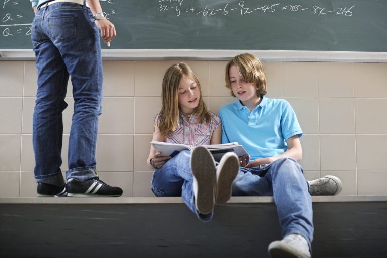 Dos niños sentados en el suelo de un aula, revisando libros. Un adulto de espaldas.